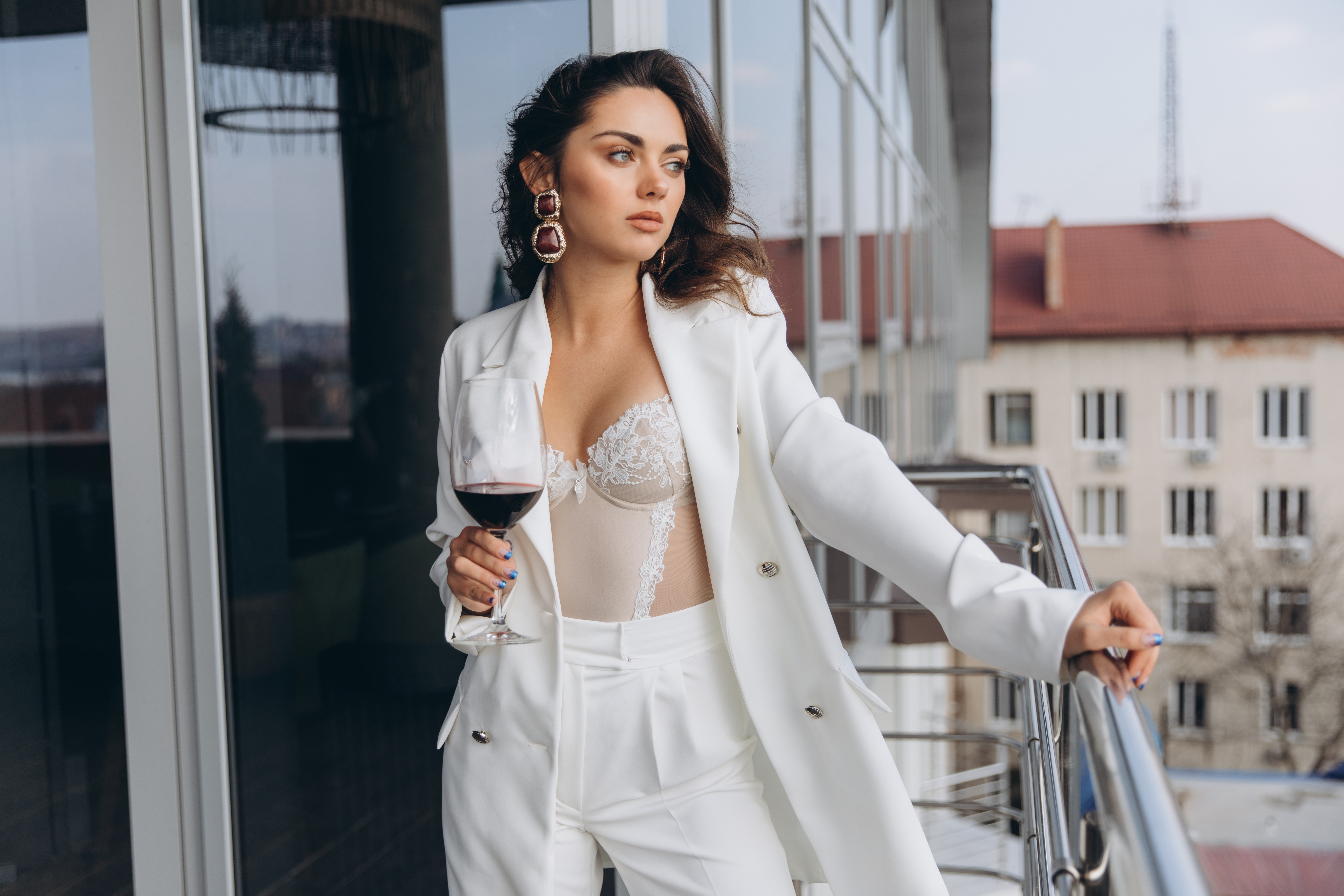 Young beautiful sexy woman in white corset and jacket relaxing at balcony with a view to the urban landscapes and drinking red wine.