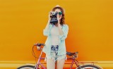 Happy young woman with film camera and bicycle on an orange background